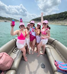 Five friends in pink swimsuits and matching pink caps cheer with canned drinks aboard a pontoon boat on a sunny lake with tree-lined rocky shoreline.