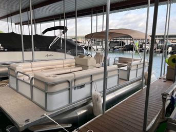 Beige pontoon boat with cushioned seating and tan bimini top moored in a covered marina slip alongside other boats, viewed from a wooden dock.