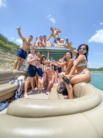 Cheerful group of young adults in swimsuits raising cans and smiling on a sunny pontoon boat at a lake with rocky shoreline and blue sky