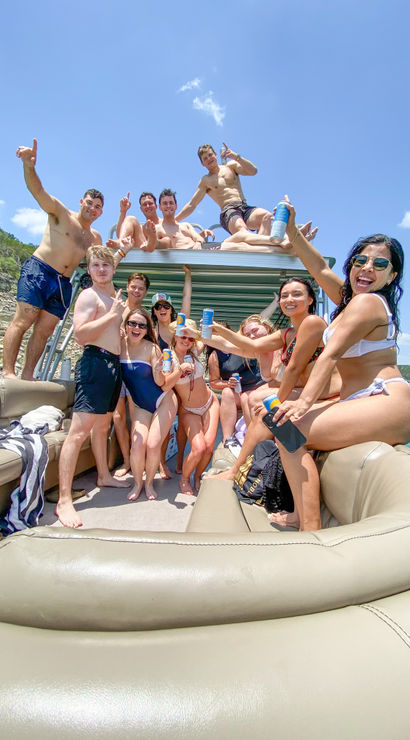 Cheerful group of young adults in swimsuits raising cans and smiling on a sunny pontoon boat at a lake with rocky shoreline and blue sky