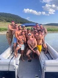 Group of shirtless men posing together on a pontoon boat at a sunny lake with wooded hills and blue sky — summer friends boating scene.