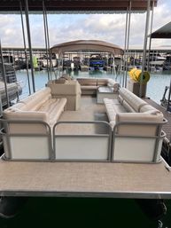 Pontoon boat with beige wraparound seating and tan canopy docked at a covered lake marina, spacious deck and calm water view.