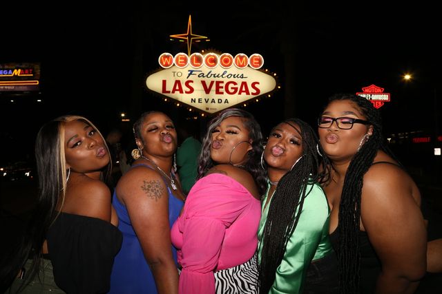 Five friends puckering for a group pose in front of the "Welcome to Fabulous Las Vegas Nevada" sign at night, colorful outfits and neon lights.