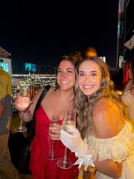 Two smiling women holding champagne flutes at a rooftop bar at night with twinkling city skyline lights, celebratory nightlife vibe.