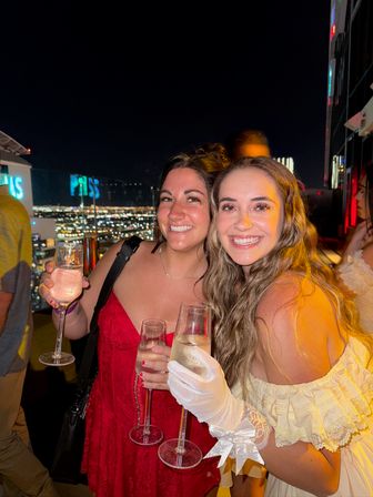 Two smiling women holding champagne flutes at a rooftop bar at night with twinkling city skyline lights, celebratory nightlife vibe.