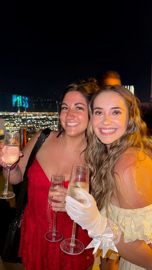 Two smiling women holding champagne flutes at a rooftop bar at night with twinkling city skyline lights, celebratory nightlife vibe.