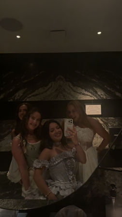 Group of four friends in formal dresses taking a mirror selfie in a dimly lit marble restroom, one holding a smartphone and smiling.