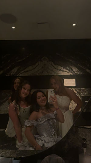 Group of four friends in formal dresses taking a mirror selfie in a dimly lit marble restroom, one holding a smartphone and smiling.