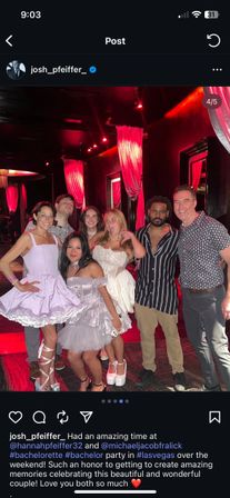 Group of seven friends posing and smiling in a red-lit Las Vegas nightclub with draped curtains; women wearing short white and lavender party dresses and platform shoes, men in casual shirts — festive bachelorette-style celebration.