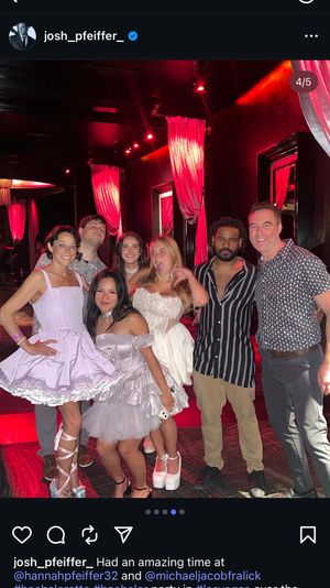 Group of seven friends posing and smiling in a red-lit Las Vegas nightclub with draped curtains; women wearing short white and lavender party dresses and platform shoes, men in casual shirts — festive bachelorette-style celebration.