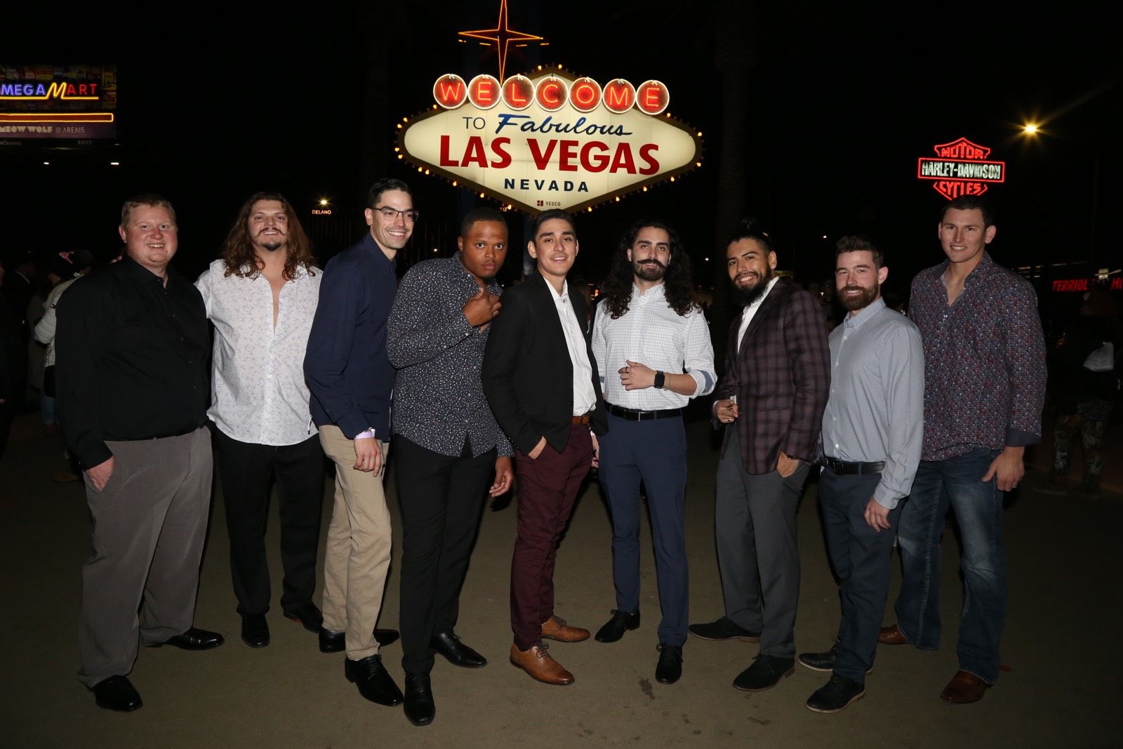 Nine men in smart-casual clothes posing at night in front of the illuminated Welcome to Fabulous Las Vegas Nevada sign, smiling for a lively group photo on the Las Vegas Strip.