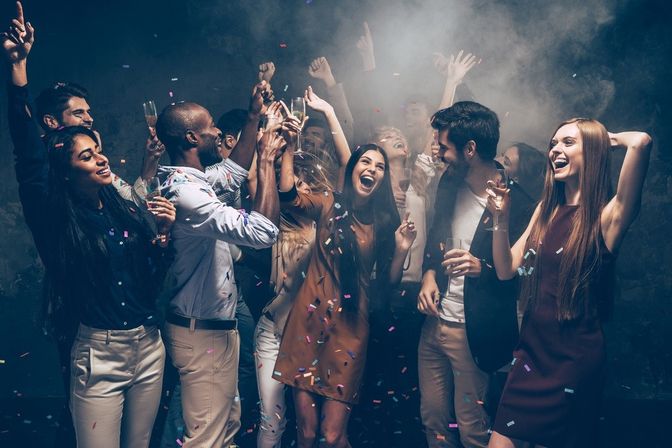 Lively indoor nightclub-style party with a diverse group of young adults cheering, dancing and toasting with champagne as confetti and smoke fill the air.