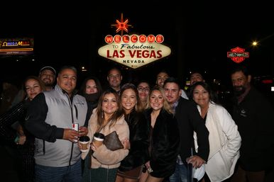 Smiling group of friends posing at night beneath the iconic Welcome to Fabulous Las Vegas neon sign on the Las Vegas Strip, holding drinks and celebrating