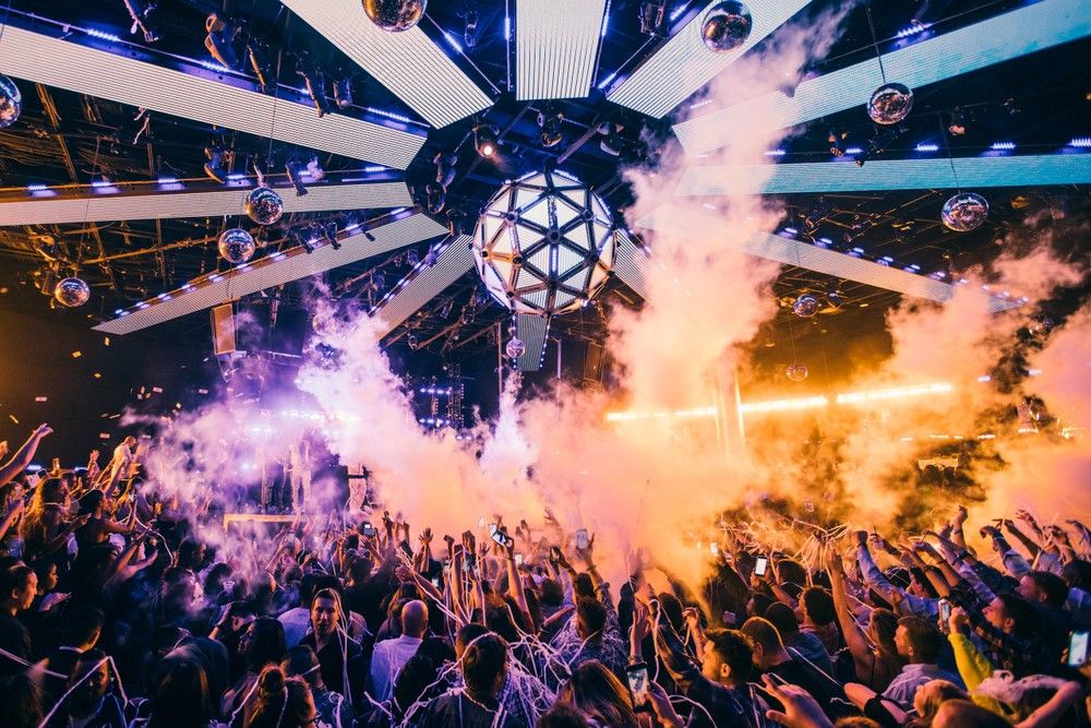 Energetic packed nightclub dance floor beneath a geometric LED disco sphere, vibrant purple-orange lights, smoke effects, confetti and raised hands