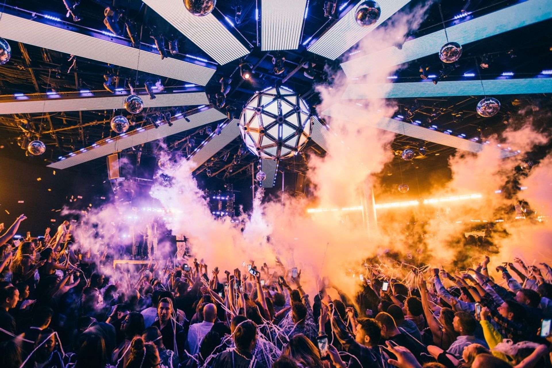 Energetic packed nightclub dance floor beneath a geometric LED disco sphere, vibrant purple-orange lights, smoke effects, confetti and raised hands