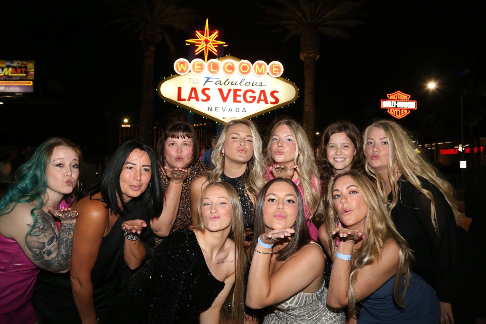Group of friends blowing kisses in front of the illuminated Welcome to Fabulous Las Vegas, Nevada sign at night, with palm trees and neon lights in the background.