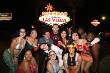 Group of friends smiling and holding red cups at night under the neon Welcome to Fabulous Las Vegas Nevada sign, festive Las Vegas Strip party scene.