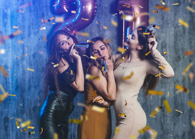 Three friends posing at an indoor birthday party, blowing party blowers under falling gold confetti with large metallic "21" balloons