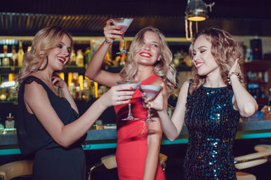 Three women in elegant dresses clink martini glasses at a stylish city bar, smiling and enjoying a fun night out in the nightlife scene.