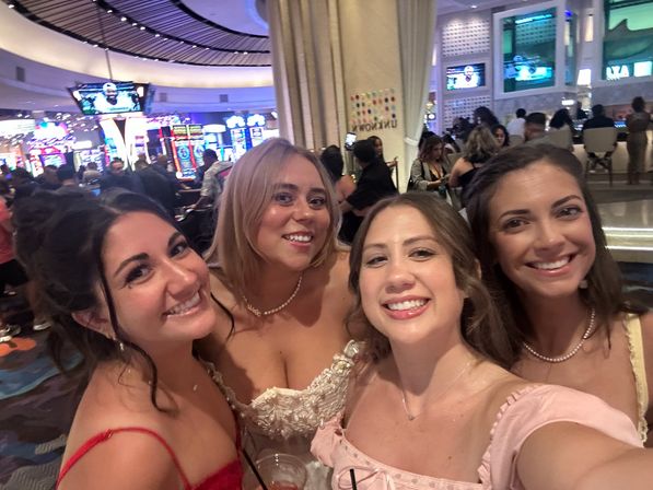 Smiling group selfie of four women dressed up on a busy casino floor with colorful slot machines and TV screens in the background