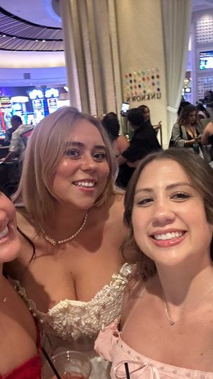 Smiling group selfie of four women dressed up on a busy casino floor with colorful slot machines and TV screens in the background