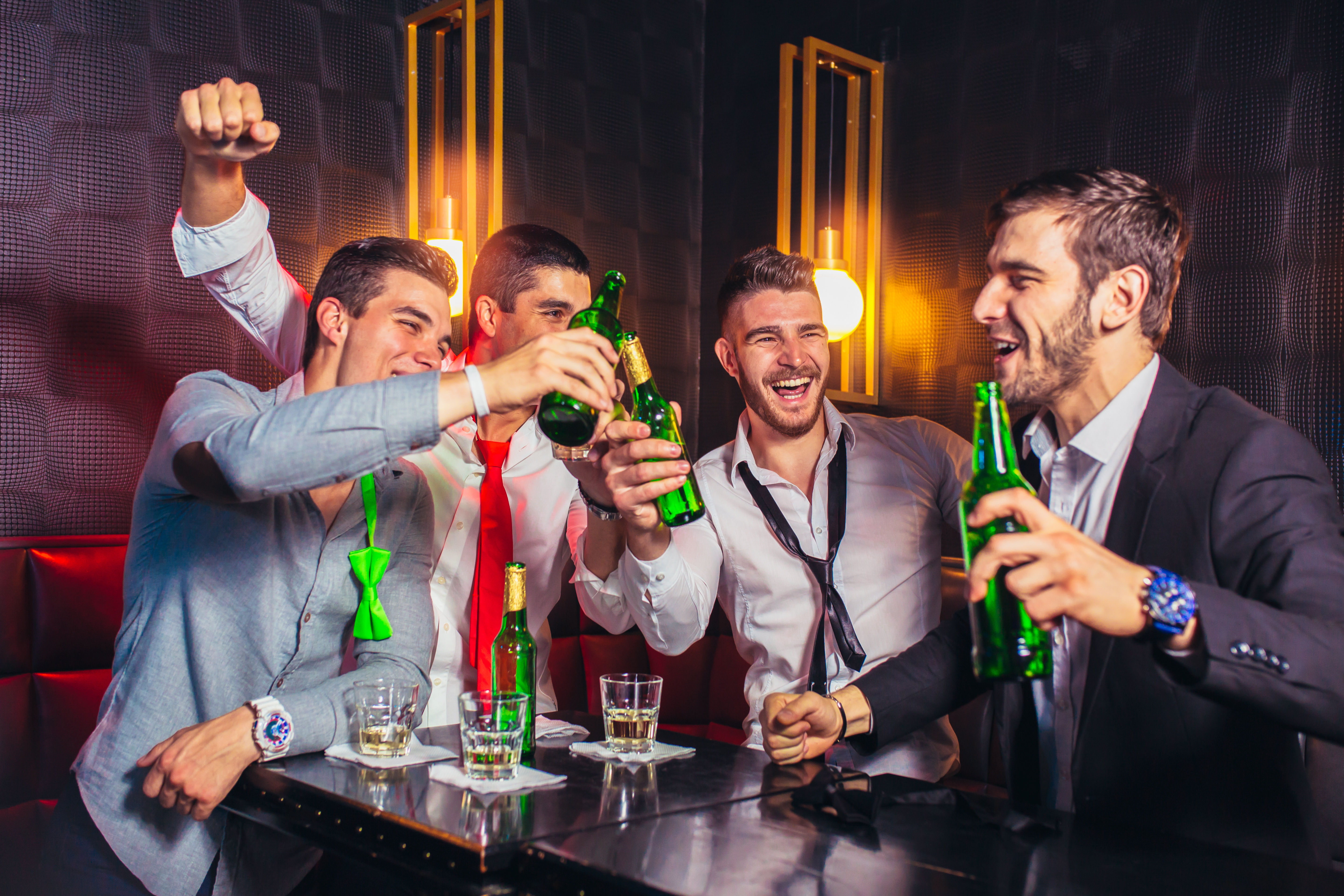 Four men in shirts and ties laughing and clinking green beer bottles in a dim modern bar booth, friends celebrating a night out in the city.