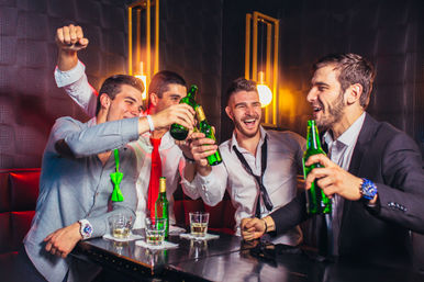 Four men in shirts and ties laughing and clinking green beer bottles in a dim modern bar booth, friends celebrating a night out in the city.