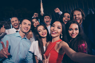 Group selfie of smiling young adults at a lively indoor nightclub party, woman in a red dress holding the camera while friends pose with peace signs, thumbs-up and cheering under warm pendant lighting — fun nightlife celebration.