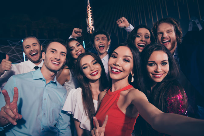 Group selfie of smiling young adults at a lively indoor nightclub party, woman in a red dress holding the camera while friends pose with peace signs, thumbs-up and cheering under warm pendant lighting — fun nightlife celebration.
