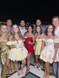 Smiling group of friends in vintage-inspired yellow and red party dresses holding champagne on a rooftop party at night with city skyline lights in the background