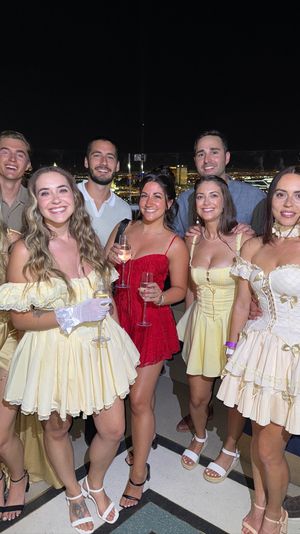 Smiling group of friends in vintage-inspired yellow and red party dresses holding champagne on a rooftop party at night with city skyline lights in the background