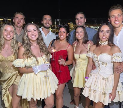 Smiling group of friends in vintage-inspired yellow and red party dresses holding champagne on a rooftop party at night with city skyline lights in the background