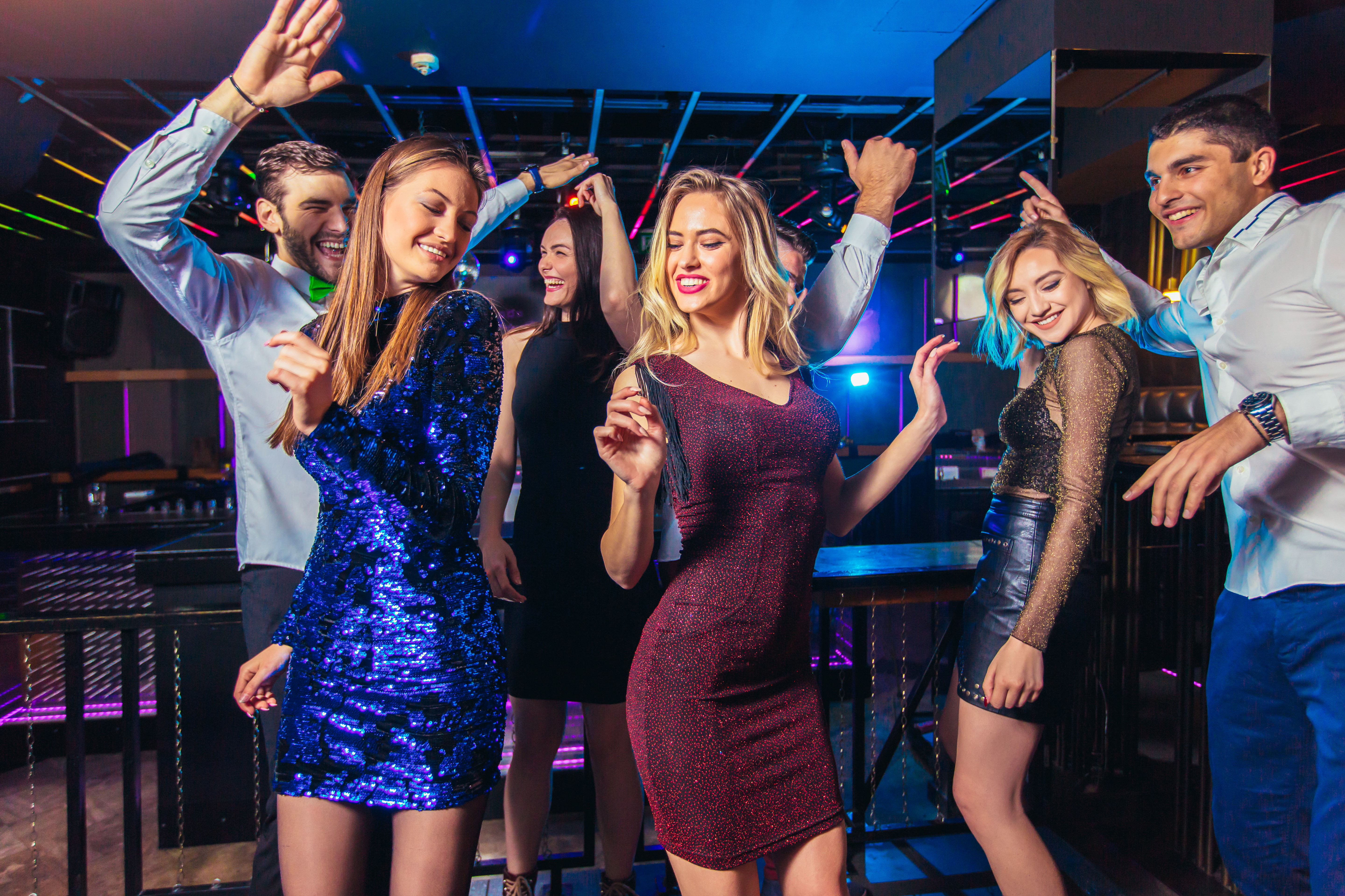 Young adults dancing on a lively downtown nightclub dance floor, women in sparkly dresses and men in shirts under colorful LED lights — energetic city nightlife party scene