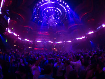 Packed nightclub dance floor under a glowing circular chandelier with purple and blue LED lighting, lively crowd dancing in a large multi-level club interior