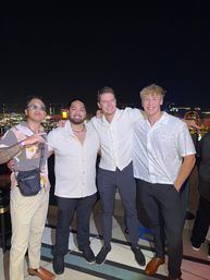 Four friends in light shirts smiling arm‑in‑arm on a rooftop terrace at night with a brightly lit city skyline and nightlife in the background