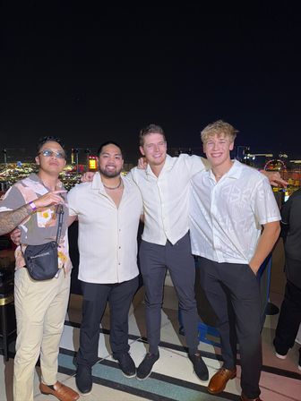 Four friends in light shirts smiling arm‑in‑arm on a rooftop terrace at night with a brightly lit city skyline and nightlife in the background