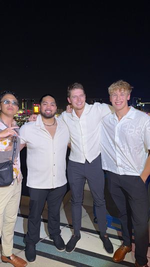 Four friends in light shirts smiling arm‑in‑arm on a rooftop terrace at night with a brightly lit city skyline and nightlife in the background