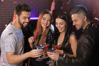 Four friends clinking colorful cocktails at a lively downtown nightclub — sequined outfits, confetti and neon lights capture an energetic city nightlife celebration.