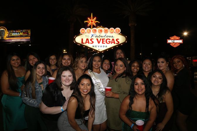 Smiling group of women posing at night in front of the illuminated Welcome to Fabulous Las Vegas, Nevada sign, holding red cups during a festive bachelorette celebration.