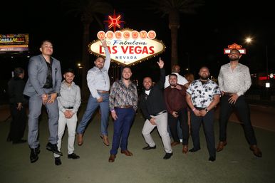 Group of men in dressy outfits posing and jumping at night in front of the illuminated Welcome to Fabulous Las Vegas, Nevada sign — a festive photo on the Las Vegas Strip.