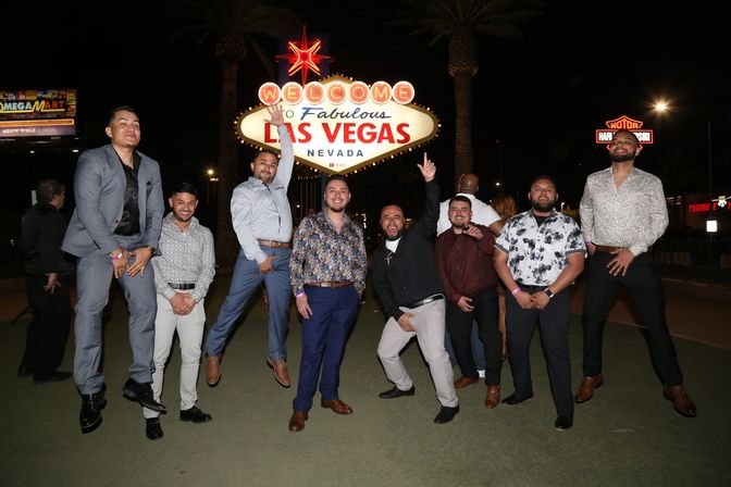 Group of men in dressy outfits posing and jumping at night in front of the illuminated Welcome to Fabulous Las Vegas, Nevada sign — a festive photo on the Las Vegas Strip.