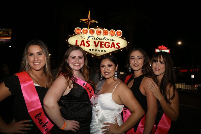 Group of five smiling women wearing bright pink bachelorette sashes, bride-to-be in a white dress, posing at night beneath the illuminated "Welcome to Fabulous Las Vegas, Nevada" sign.