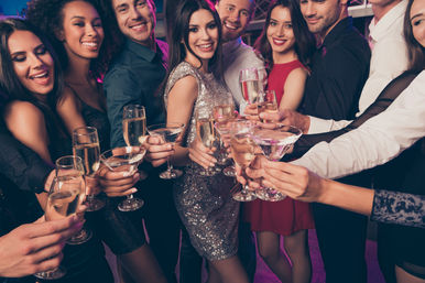 Smiling group of young adults in evening wear raising champagne flutes and cocktails for a celebratory toast in a crowded nightclub with purple lighting