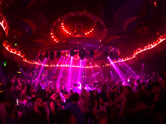 Packed nightclub dance floor under a large circular LED chandelier, magenta and red spotlights and laser beams cutting through haze as a lively crowd dances in a vibrant nightlife scene.