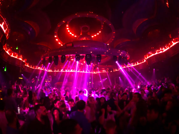 Packed nightclub dance floor under a large circular LED chandelier, magenta and red spotlights and laser beams cutting through haze as a lively crowd dances in a vibrant nightlife scene.