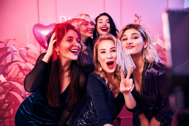 Five friends in sparkly dresses pose for a selfie in a pink-lit photo booth at a bachelorette party, wearing veils, smiling and flashing a peace sign amid balloons and streamers.