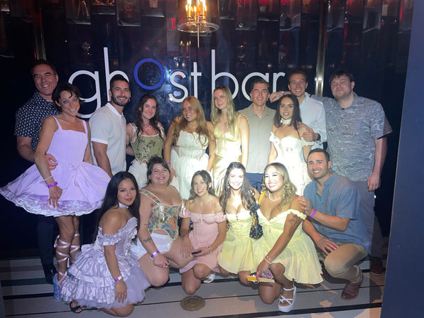 Large group of friends smiling for a celebratory group photo in a dim nightclub with a lit wall sign, many wearing pastel corset dresses, playful skirts and casual shirts — lively nightlife party scene.