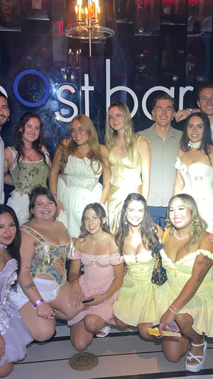 Large group of friends smiling for a celebratory group photo in a dim nightclub with a lit wall sign, many wearing pastel corset dresses, playful skirts and casual shirts — lively nightlife party scene.