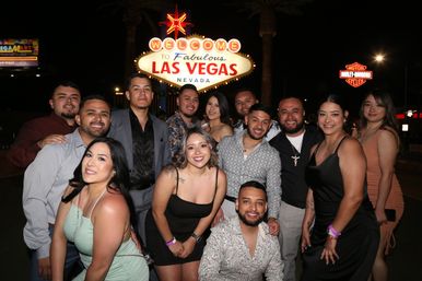 Smiling group of adults posing at night in front of the illuminated Welcome to Fabulous Las Vegas Nevada sign on the Strip, palm trees and city lights in the background