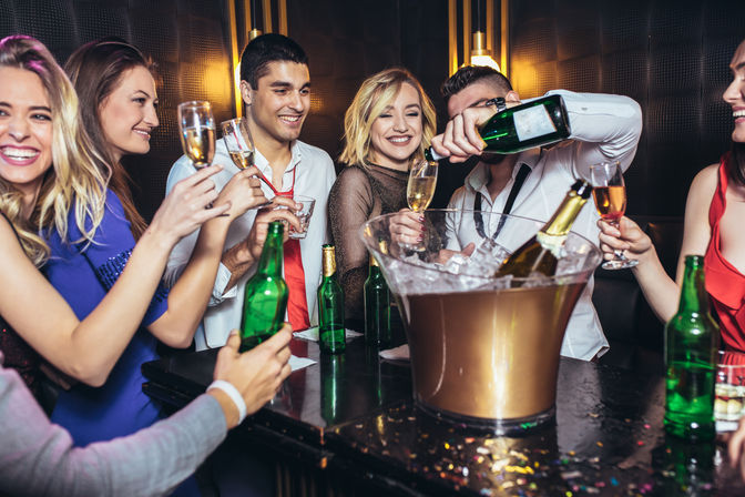 Lively nightclub bar scene with a group of adults toasting with champagne flutes while a server pours from a bottle beside an ice-filled champagne bucket — festive party atmosphere.